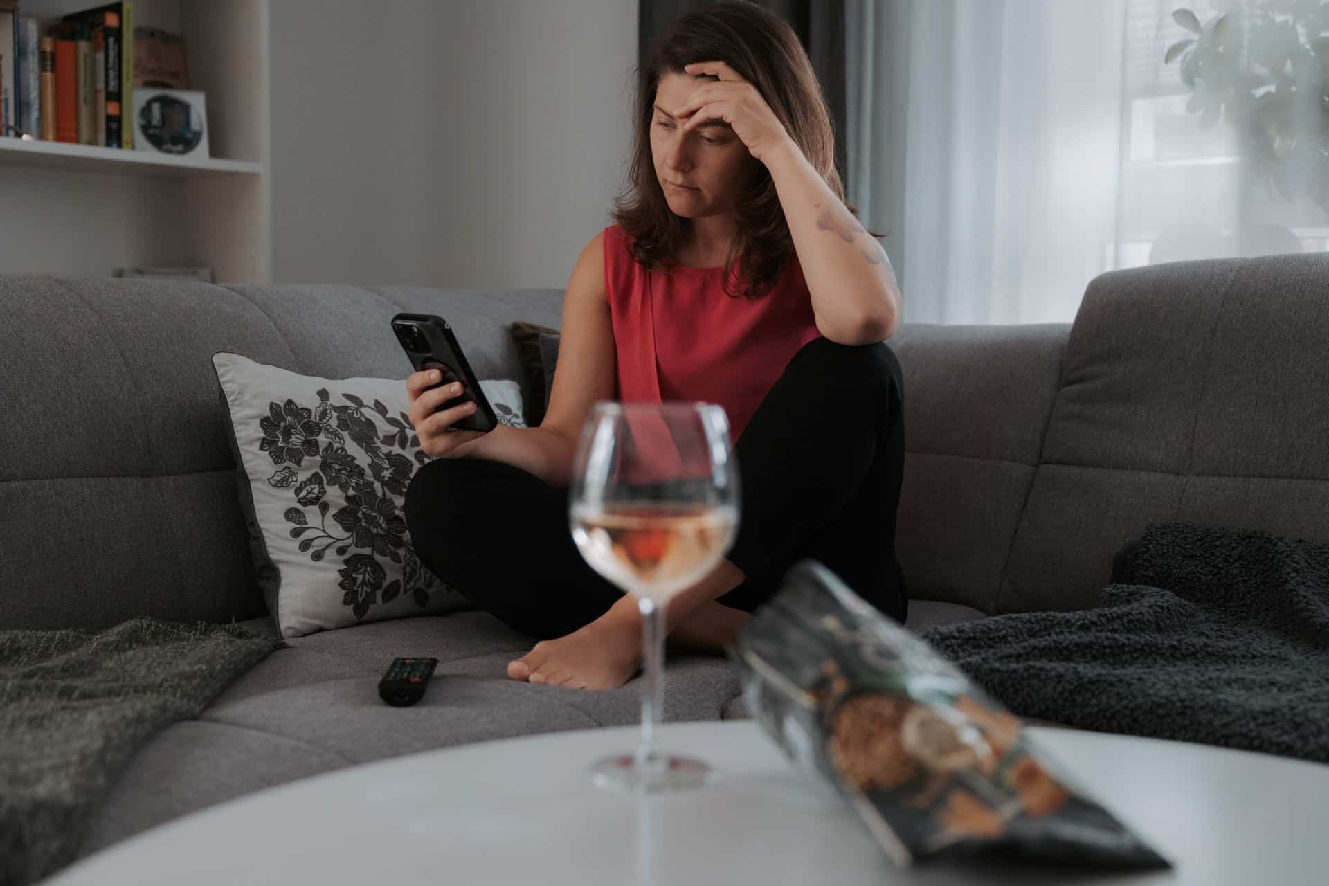 Frau sitzt allein auf dem Sofa, hält ihr Handy und fasst sich besorgt an die Stirn