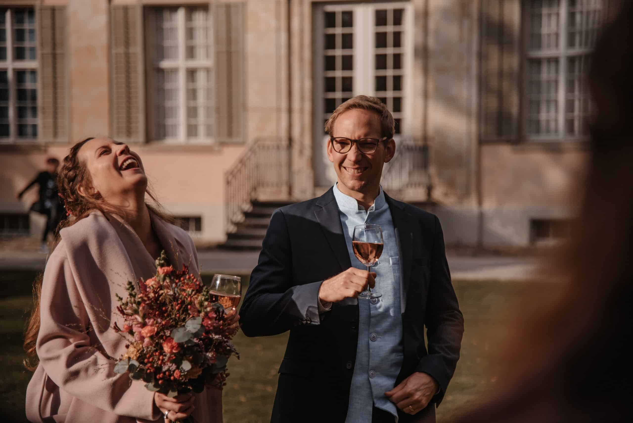 Lachendes Paar mit Blumenstrauß und Gläsern bei einer Feier vor einem hellen Herrenhaus